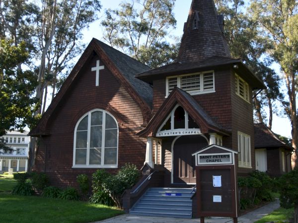 Image of a quaint brown church with a steep-roofed steeple, gothic windows, and a signboard in front, surrounded by trees and green grass.