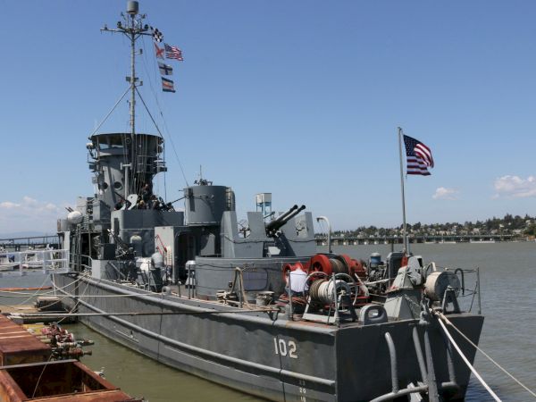 A military ship with the number 102 is docked at a pier, displaying an American flag. The background shows water and a distant shoreline.