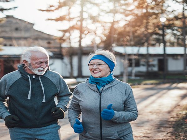 Two elderly people jog together on a tree-lined path with smiles on their faces, enjoying a sunny day outdoors.