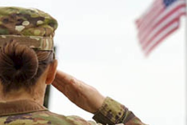 A soldier in camouflage uniform is saluting an American flag, seen from behind, with a patch on the arm of the uniform clearly visible.