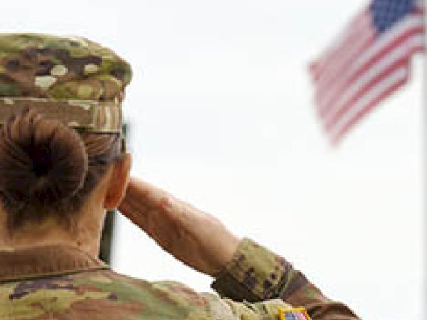 A soldier in camouflage uniform is saluting an American flag, seen from behind, with a patch on the arm of the uniform clearly visible.
