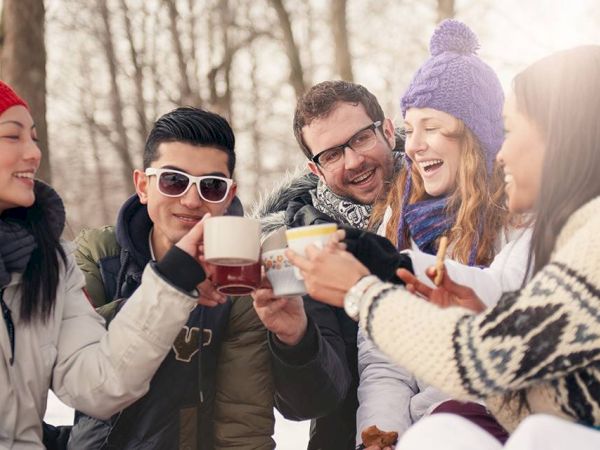 A group of friends wearing winter clothes is outdoors, smiling, and toasting with mugs in a snowy setting.