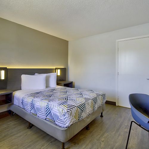 A modern hotel room features a double bed with patterned bedding, a desk with a chair, and built-in wall lighting on a wooden floor.