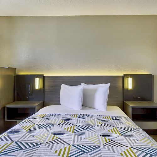 A hotel room with a neatly made bed, patterned bedspread, two pillows, nightstands with lamps, microwave, and mini-fridge in the corner.