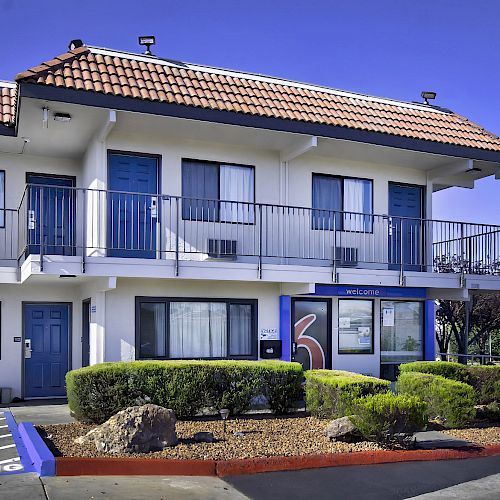 The image shows a two-story motel with blue doors and a red-tiled roof, featuring neatly trimmed bushes and a 