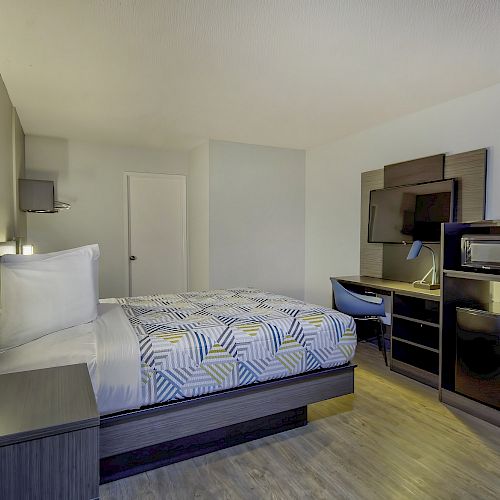 A modern hotel room with a double bed, flat-screen TV, desk, chair, microwave, mini-fridge, and wood flooring, all with a minimalist design.