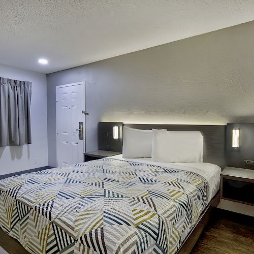 The image shows a minimalist hotel room with a double bed, modern patterned bedspread, nightstands, wall lamps, curtains, and a door.