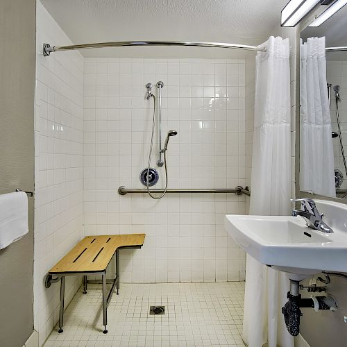 This is a bathroom with a sink, a shower area equipped with a bench and handrails, and towels folded and stacked on a shelf.