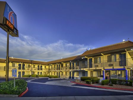 The image shows the exterior of a Motel 6 with a sign and a parking lot in the front, surrounded by bushes and a multi-story building.