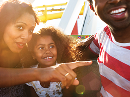 A woman, a man, and a child are enjoying a fun moment outdoors, possibly at an amusement park, with bright sunlight in the background.