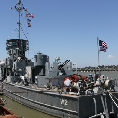 A military ship marked with the number 102 is docked at a pier. It flies an American flag, and various equipment is visible on deck.