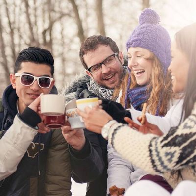 A group of friends in winter clothing are outdoors, toasting with mugs and smiling, with a snowy background and sunlight shining through the trees.