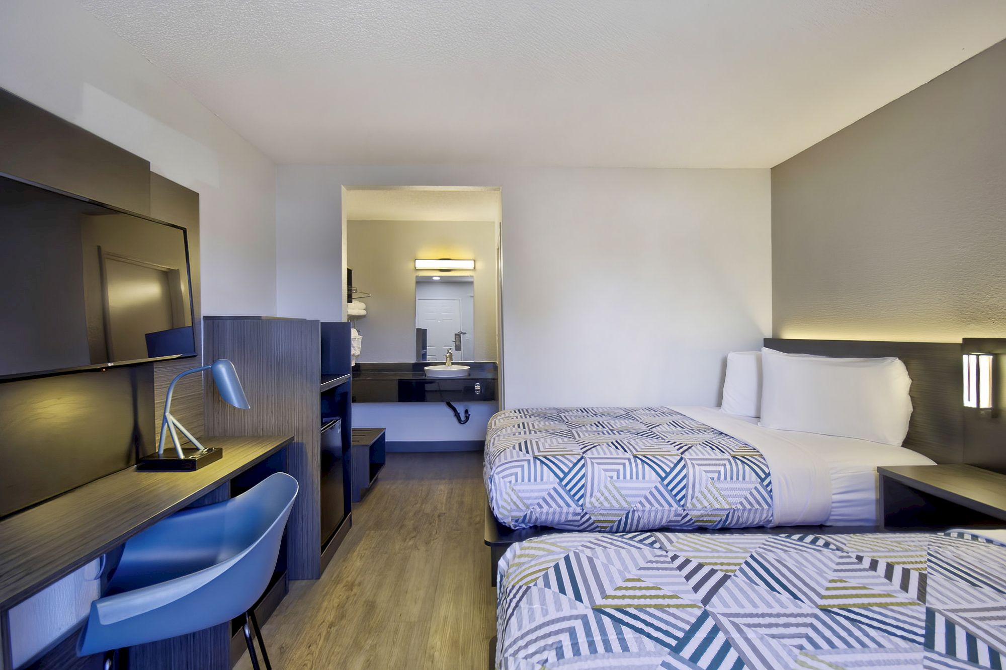 A modern hotel room with two beds, a desk with a chair and lamp, a wall-mounted TV, and a sink area visible through a partition.