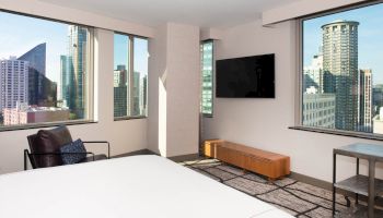 A modern hotel room with large windows, a bed, a wall-mounted TV, a chair, a small table, and a cityscape view with tall buildings outside.