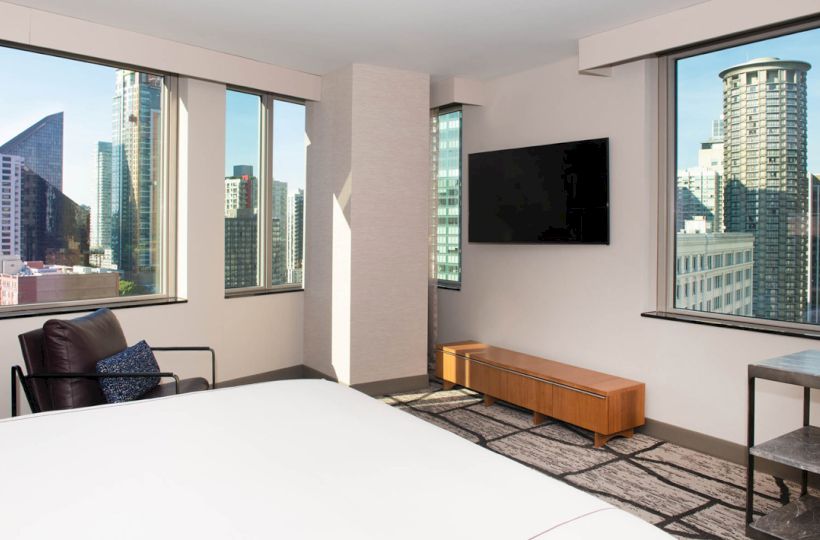 A modern hotel room with large windows, a bed, a wall-mounted TV, a chair, a small table, and a cityscape view with tall buildings outside.