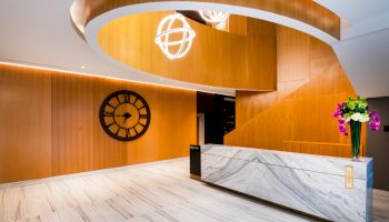 A modern lobby with a marble reception desk, a large wall clock, a curved wooden staircase, and contemporary light fixtures with a floral arrangement.