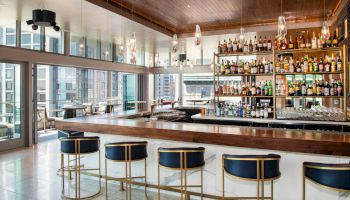 A modern bar with a wooden counter, five high chairs, and shelves stocked with various bottles of alcohol. Large windows offer cityscape views.