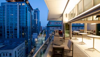 A modern balcony overlooking a city street with tall buildings, outdoor seating, and tables, illuminated warmly during twilight.