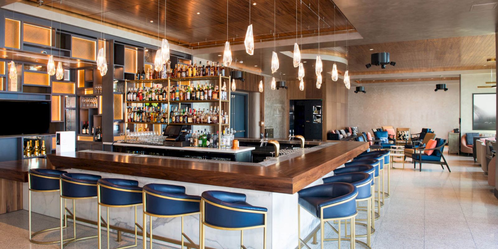 The image shows a modern bar with blue and gold barstools, a wooden counter, hanging light fixtures, and shelves stocked with various bottles.