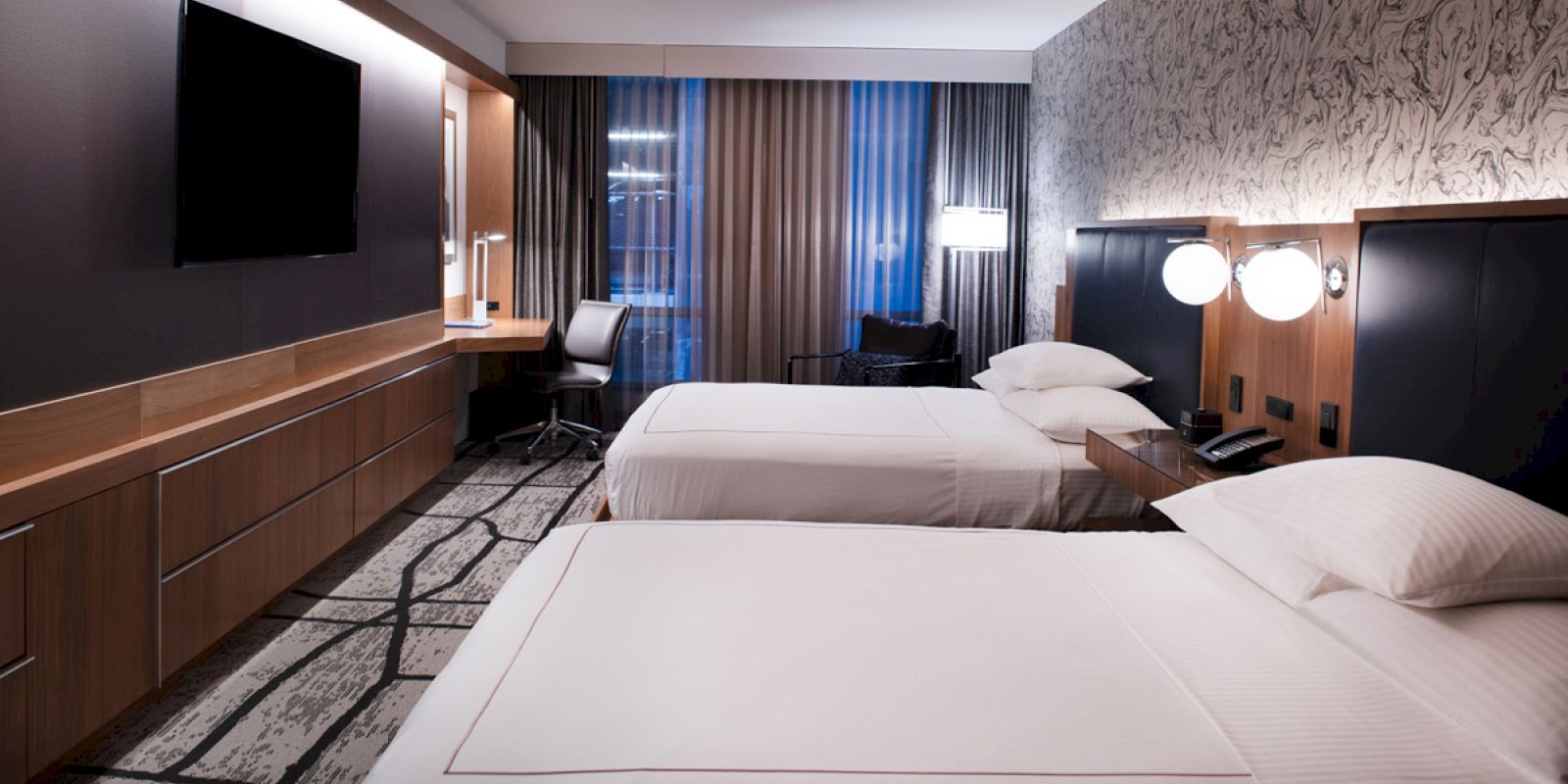 A modern hotel room with two neatly made beds, a desk by the window, a flat-screen TV on the wall, and stylish interior decor.