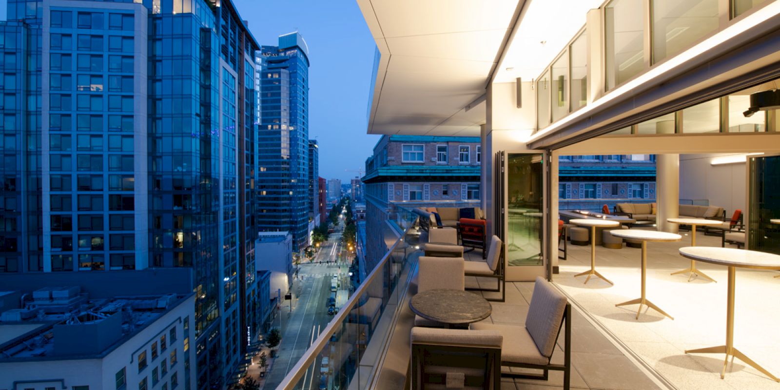 A modern balcony with chairs and tables overlooks a city street lined with tall buildings as evening sets in and lights illuminate the scene.