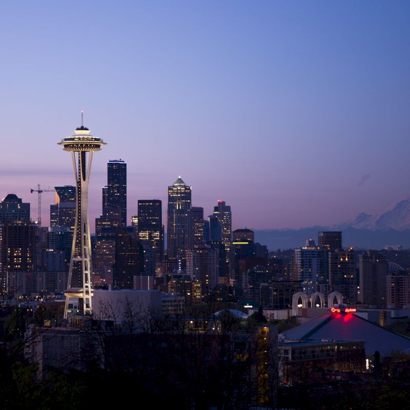 The image shows the Seattle skyline at dusk, featuring the Space Needle, city buildings, and Mount Rainier in the background against a twilight sky.