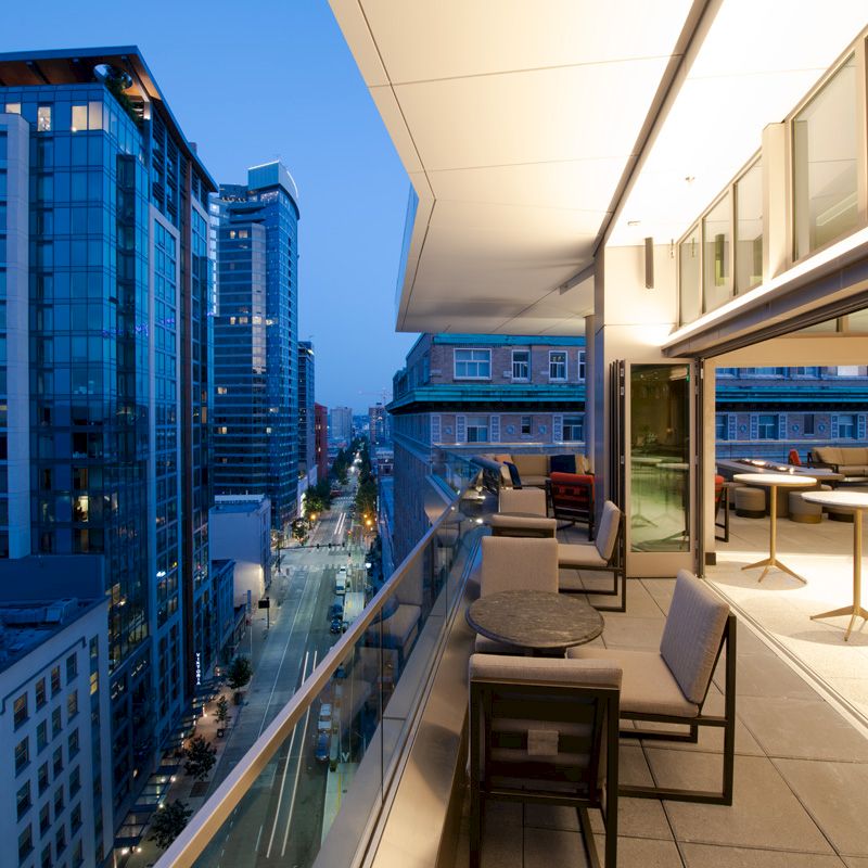 A modern cityscape with tall buildings is seen from a rooftop terrace, featuring outdoor seating and tables.