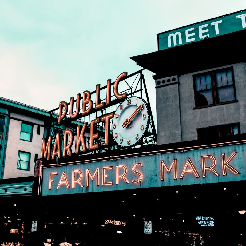 The image shows a vibrant neon sign for a "Public Market" with a clock, and another sign beneath it reads "Farmers Market" on historic buildings.