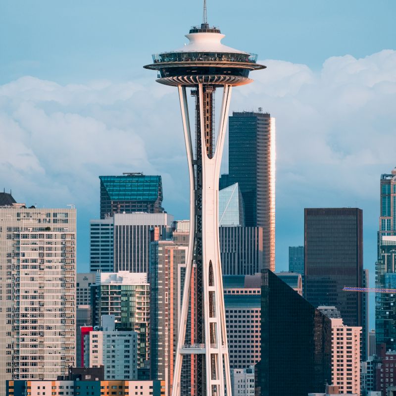 The image features the Space Needle, a prominent observation tower, surrounded by downtown Seattle's modern skyscrapers against a backdrop of clouds.