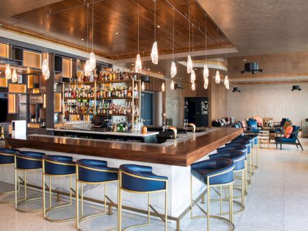 This image shows a modern bar with a central counter, bar stools, hanging lights, and a well-stocked liquor shelf, with seating areas in the background.