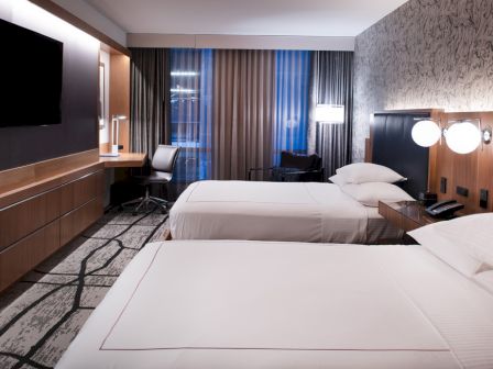 The image shows a modern hotel room with two neatly made beds, a desk by the window, a TV on the wall, and contemporary decor.