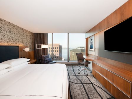 A modern hotel room with a large bed, desk, chair, TV, and a window offering a city view.