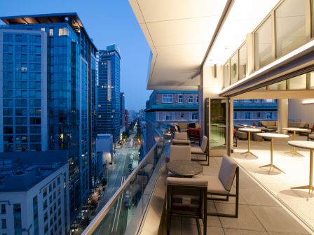 A rooftop balcony overlooks a city street lined with modern high-rise buildings, featuring outdoor seating areas and large windows.