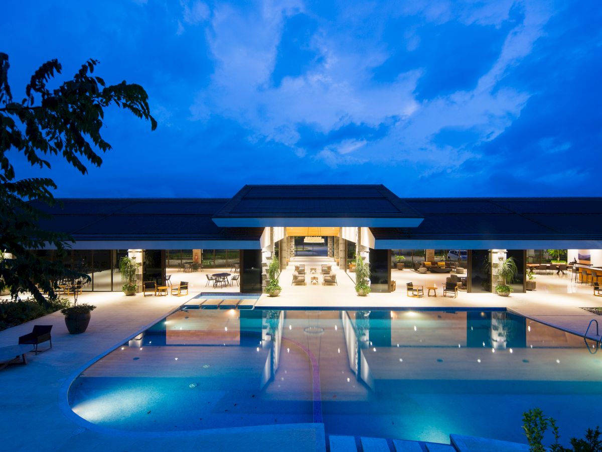 A modern, well-lit building with a large pool in front, surrounded by lounge chairs and greenery, under a twilight sky.