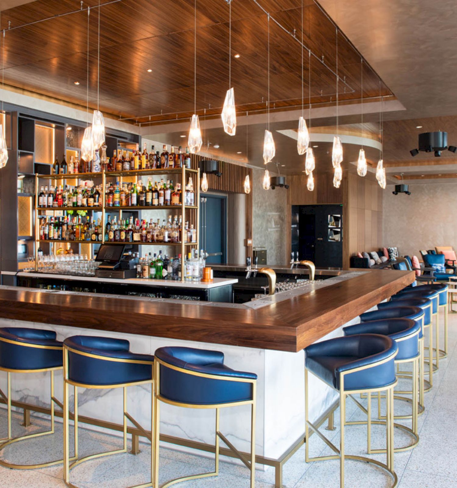 The image shows a modern bar with a sleek design. It has a row of blue barstools, a well-stocked bar, hanging lights, and a lounge area with seating.