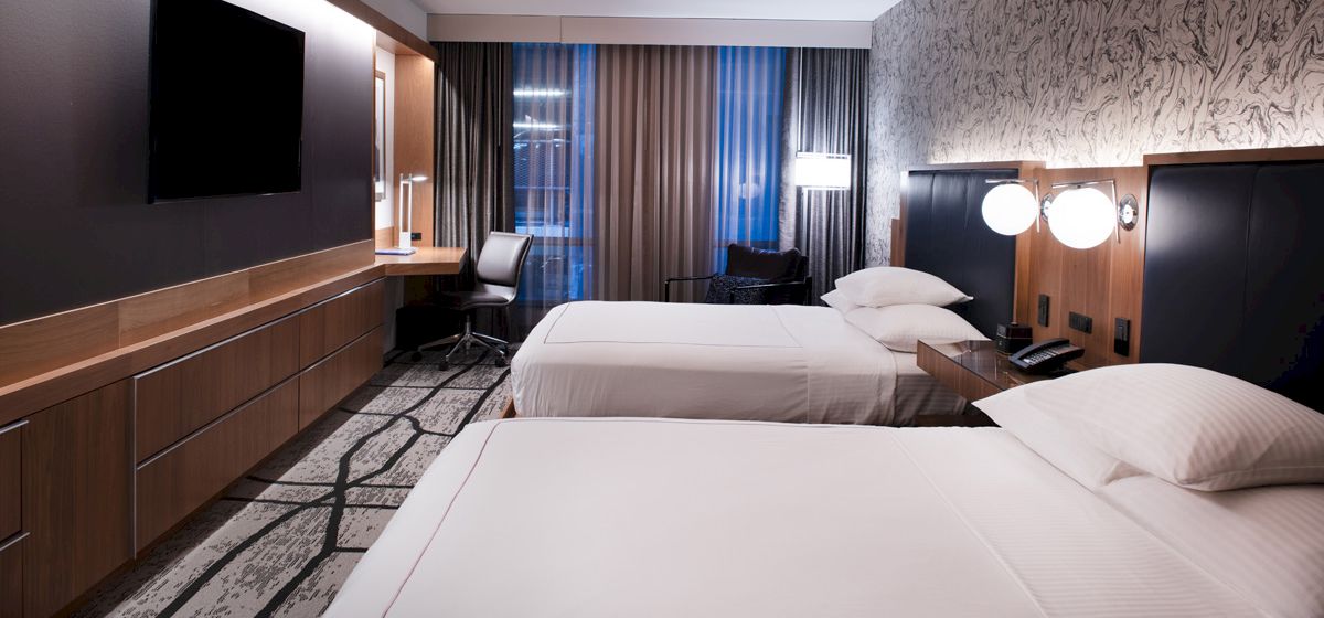 A modern hotel room with two neatly made beds, a flat-screen TV, a work desk, ambient lighting, and a large window with curtains.