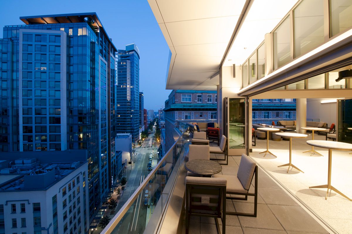 A modern urban balcony at dusk with outdoor seating overlooks a city street and surrounding high-rise buildings illuminated by streetlights.