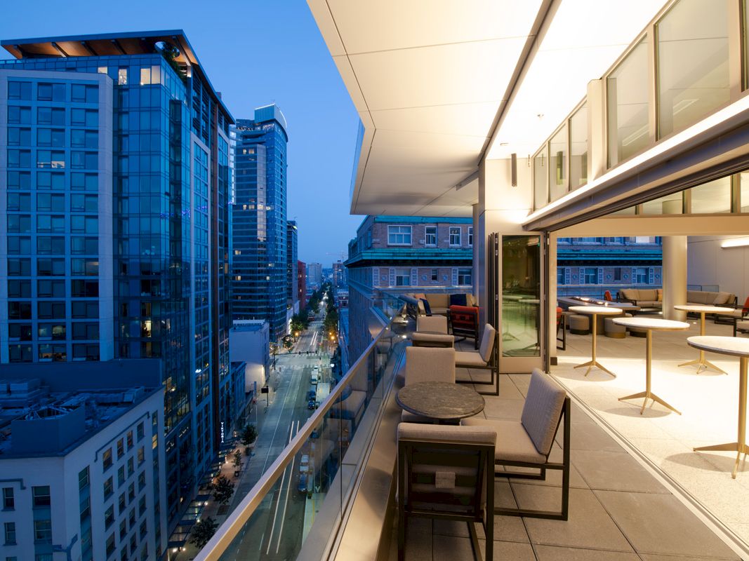 A modern urban balcony at dusk with outdoor seating overlooks a city street and surrounding high-rise buildings illuminated by streetlights.
