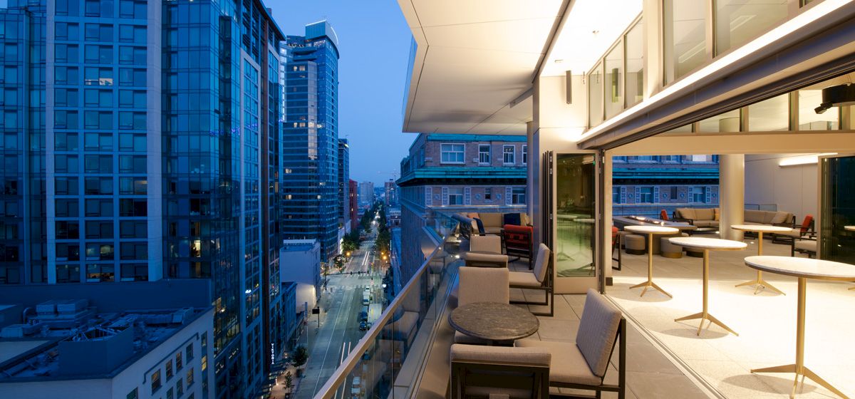 A modern urban balcony at dusk with outdoor seating overlooks a city street and surrounding high-rise buildings illuminated by streetlights.