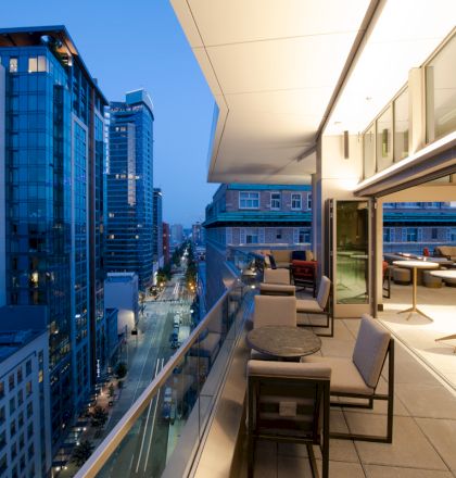 A modern cityscape viewed from a high-rise balcony at twilight, with outdoor seating areas and tall buildings lining a busy street.