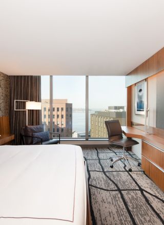 A modern hotel room with a large bed, desk, chair, and a big-screen TV, featuring a city view through floor-to-ceiling windows.