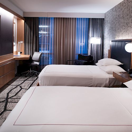 The image shows a modern hotel room with two neatly made beds, a wall-mounted TV, a desk, chair, and stylish decor.