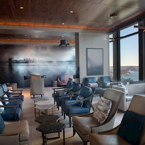 The image shows a modern lounge with leather chairs, tables, wall art, and large windows offering a view of a waterfront and Ferris wheel.