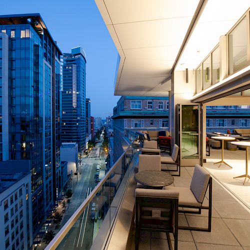 This image shows a modern balcony with seating and tables overlooking a city street lined with buildings in the evening light.