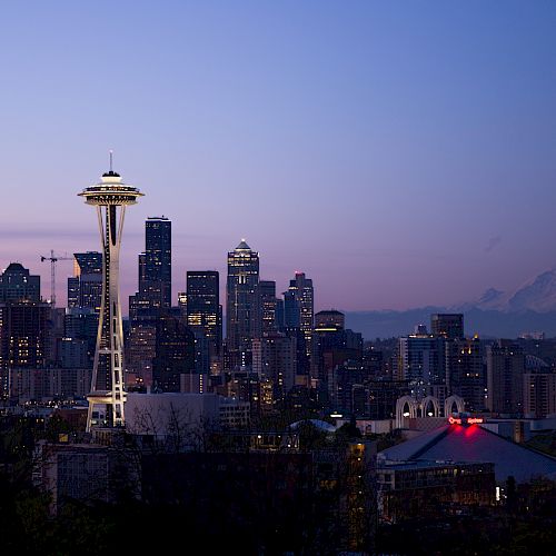 The image shows the Seattle skyline at dusk, featuring the iconic Space Needle and Mount Rainier in the background, with city lights beginning to twinkle.