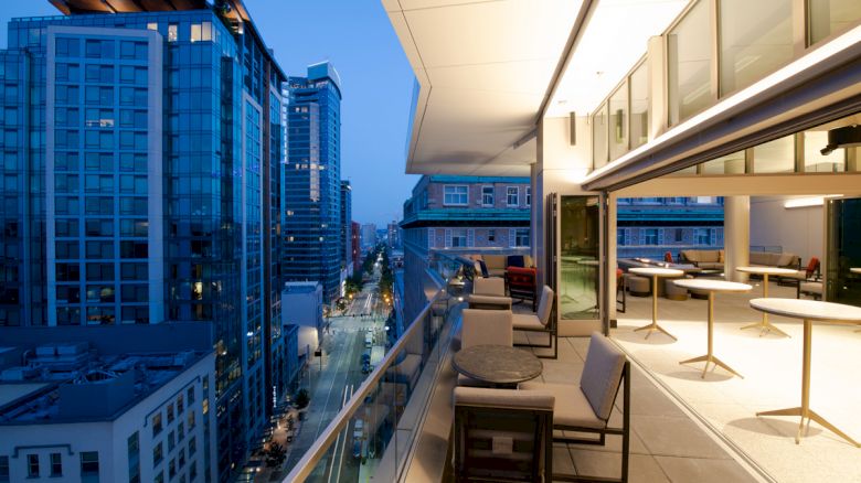 A modern cityscape at dusk with high-rise buildings, viewed from a stylish rooftop terrace with seating and tables, along an illuminated street.