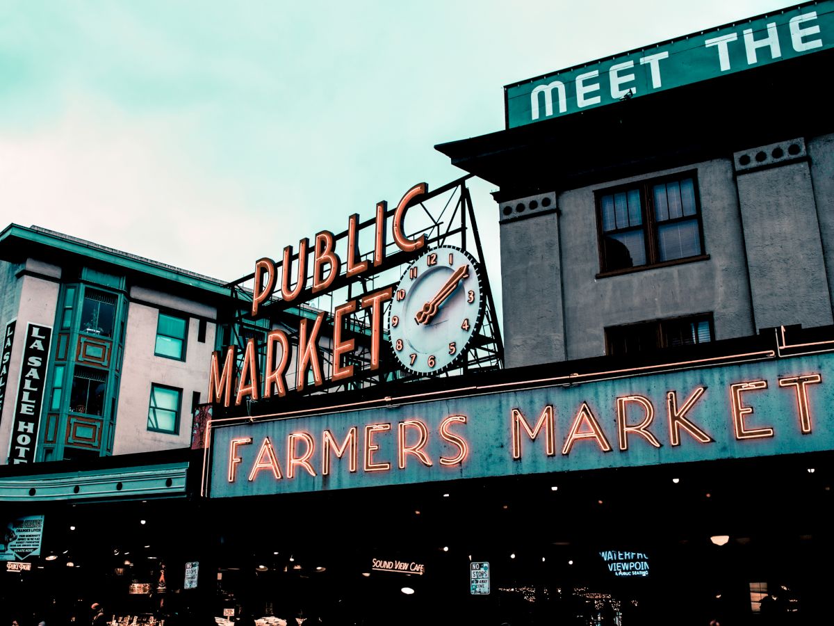 The image shows a public market with prominent neon signs stating 'Public Market' and 'Farmers Market' against a backdrop of buildings.