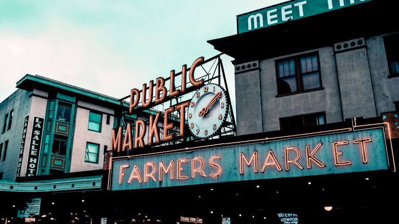 The image shows a public market with prominent neon signs stating 'Public Market' and 'Farmers Market' against a backdrop of buildings.