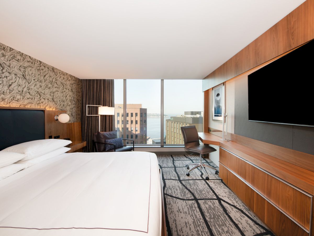 A modern hotel room features a large bed, desk, chair, wall-mounted TV, and a scenic city view through a large window.