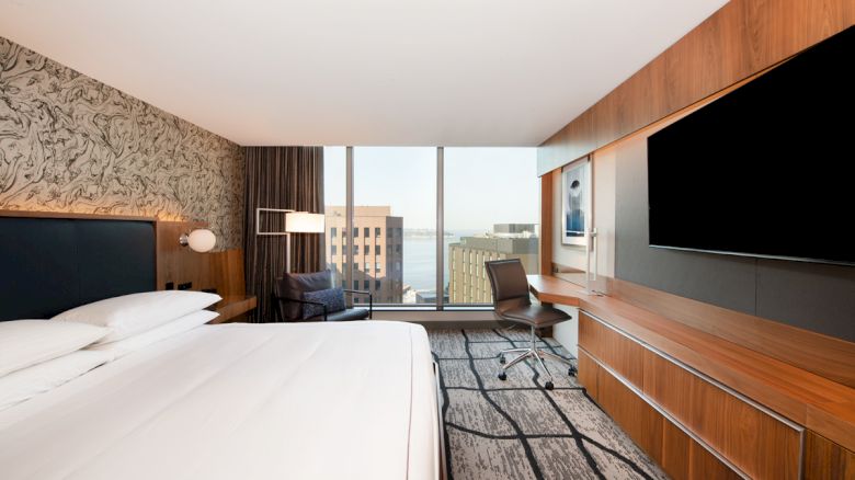 A modern hotel room features a large bed, desk, chair, wall-mounted TV, and a scenic city view through a large window.